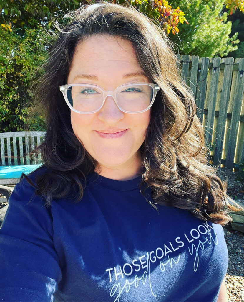 Michelle, a white woman with brown hair, faces the camera with a smile. She wears glasses with clear frames and a shirt that says, "Those Goals Look Good On You."