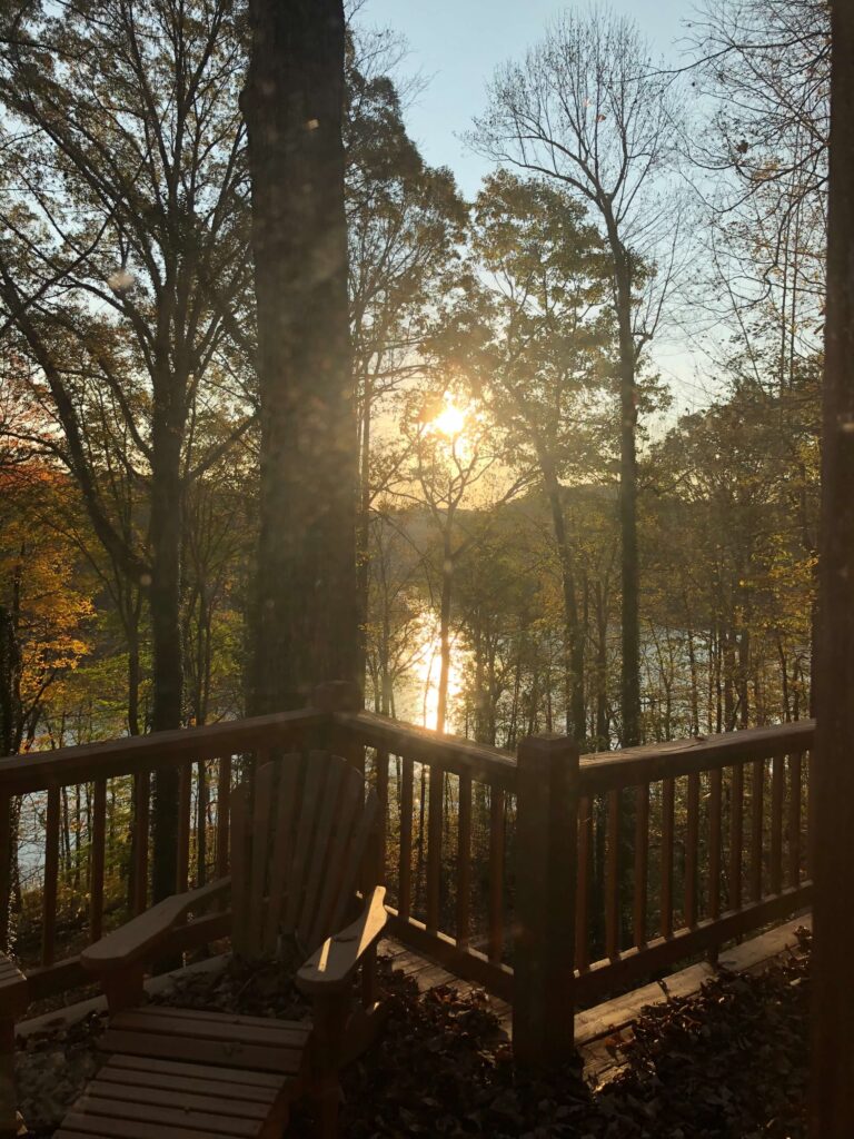 The sun shines over a lake. There are hills in the background, and a deck with an Adirondack chair in the foreground. Large trees stand between the deck and the lake's edge.