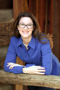 Michelle, a white woman with brown hair, is leaning against a log railing. She is wearing glasses and a blue collared shirt.