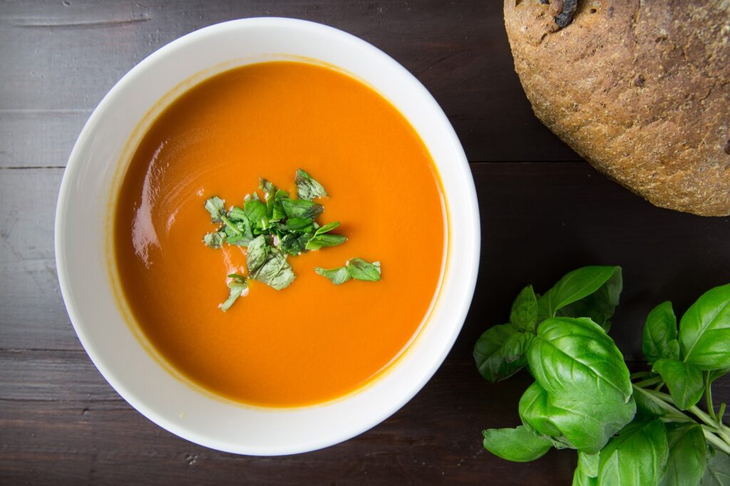 A bowl of tomato soup garnished with basil rests beside a loaf of crusty bread on a dark wooden table. Find a sprig of basil in the bottom right corner.