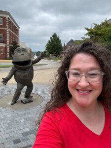 Michelle is smiling and wearing a red shirt with the Hilltopper mascot statue, "Big Red" in the background over her shoulder.